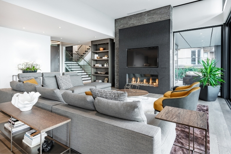 Modern living room with a large gray sectional sofa, built-in shelves, a fireplace under a wall-mounted TV, two mustard chairs, a large window, and green indoor plants, decorated in neutral tones.