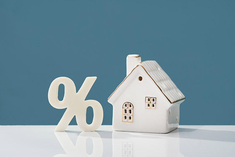 A small ceramic house and a large white percentage symbol are placed side by side on a reflective surface, against a plain blue background.